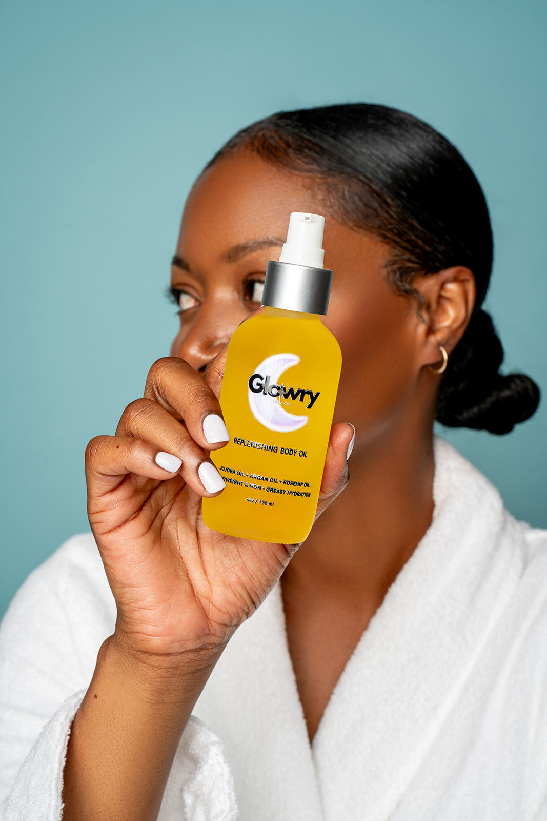 Woman in a white robe smiling and holding a bottle of Glowry Skin Co. Replenishing Body Oil for sensitive skin.