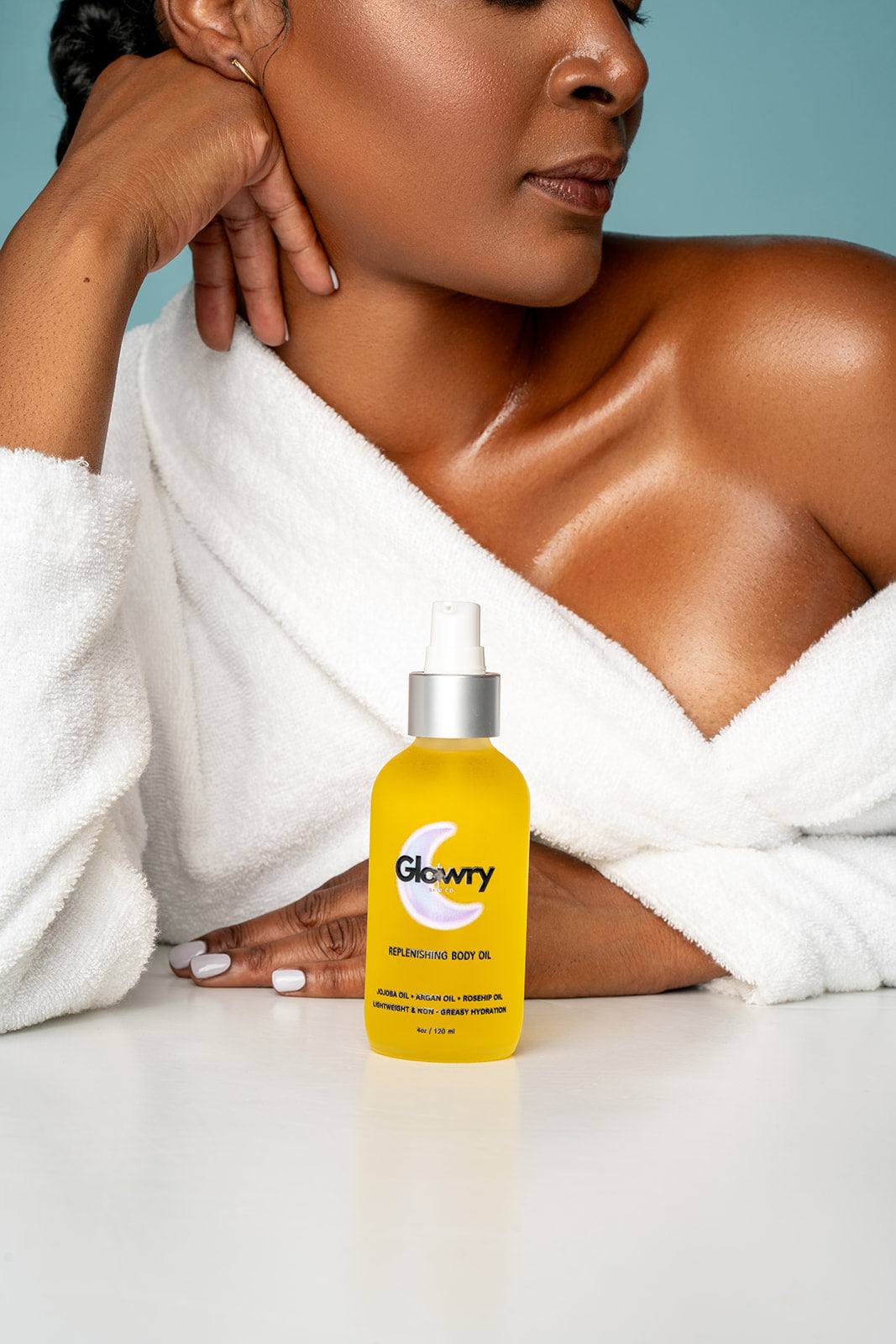 Woman in a white robe with glowing, hydrated skin on her chest and shoulders, sitting with a bottle of Glowry Replenishing Body Oil.